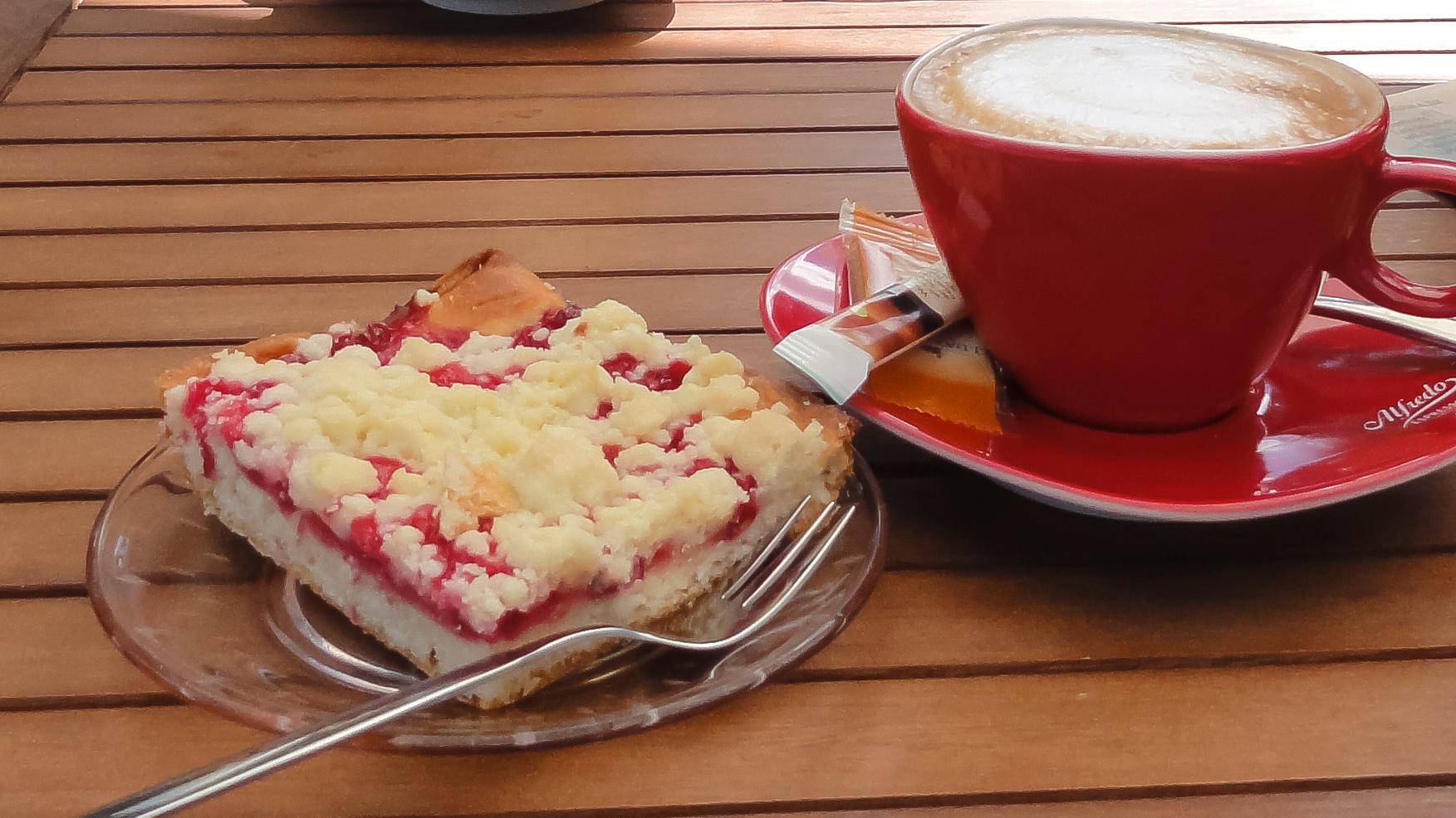 Man sieht ein Stück Streuselkuchen auf einem roten Teller, rechts daneben eine rote Tasse mit Kaffee auf einer Untertasse