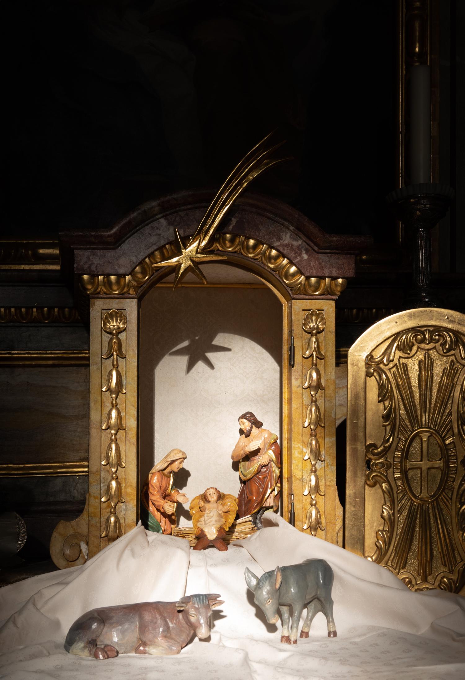 Man sieht eine goldumrahmte Krippe mit Maria, Josef und Jesuskind, davor Ochs und Esel auf einem weißen Tuch