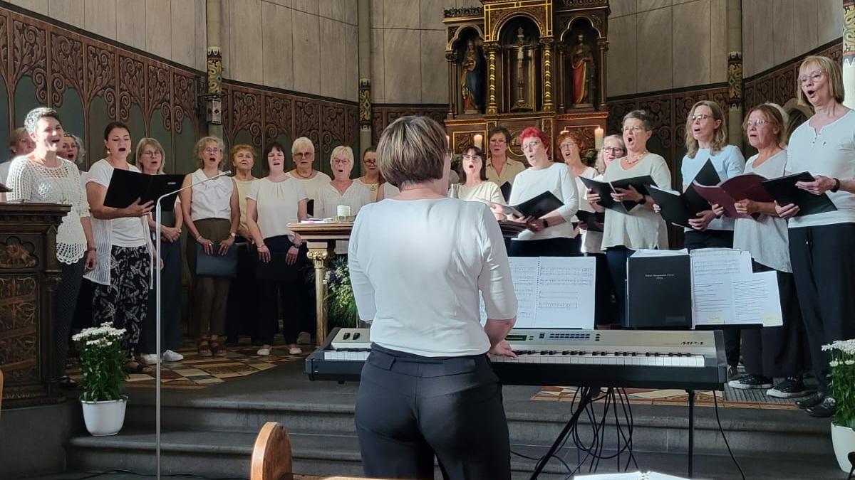 Man sieht Frauen mit Notenbüchern in weißen Blusen und schwarzen Hosen in der Apsis der Kirche in Rüber singen, davor die Chorleiterin