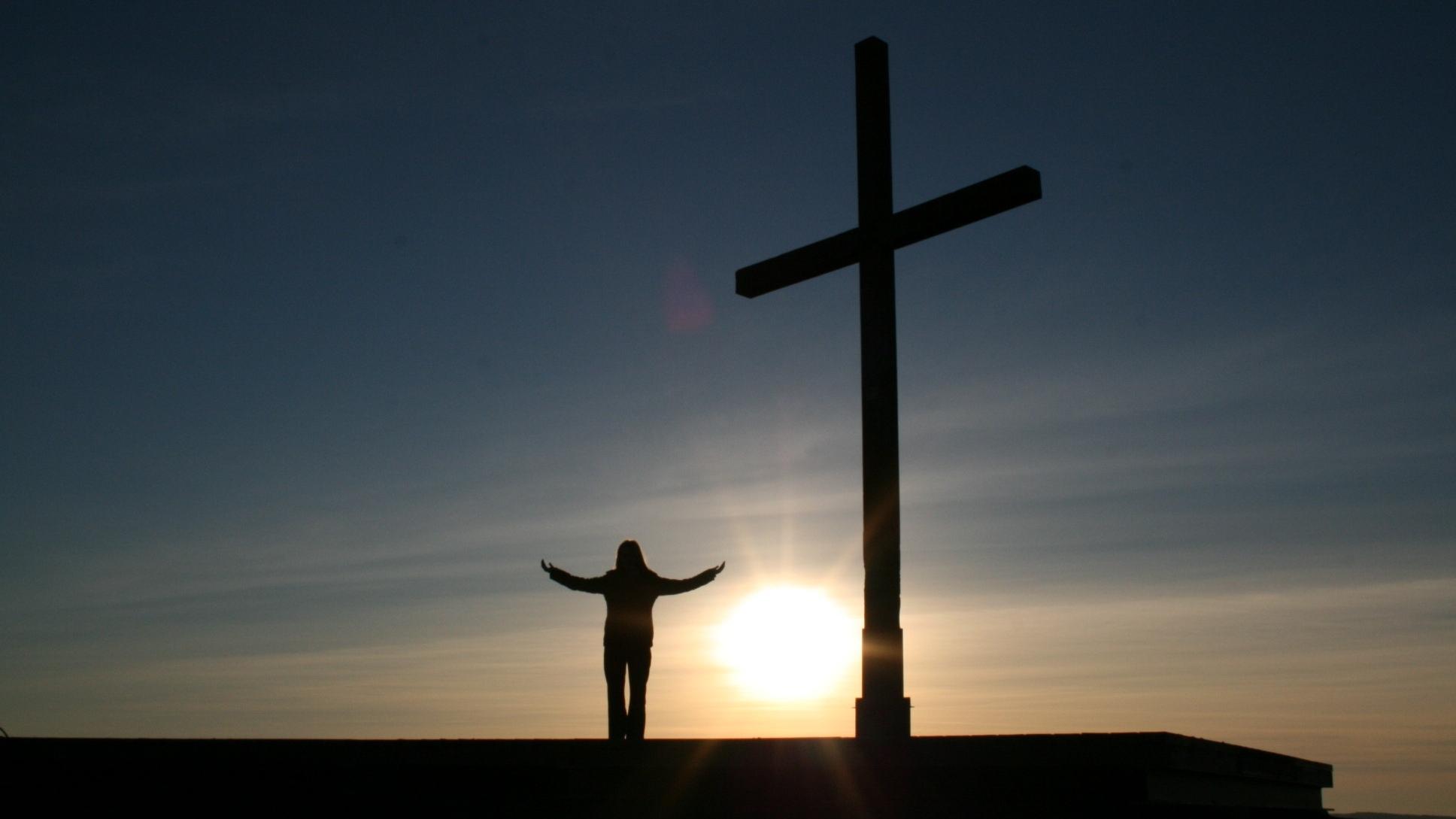 Im Vordergrund links ein Kreuz vor untergehender Abendsonne - rechts ein Mensch im Gebet mit ausgebreiteten Händen