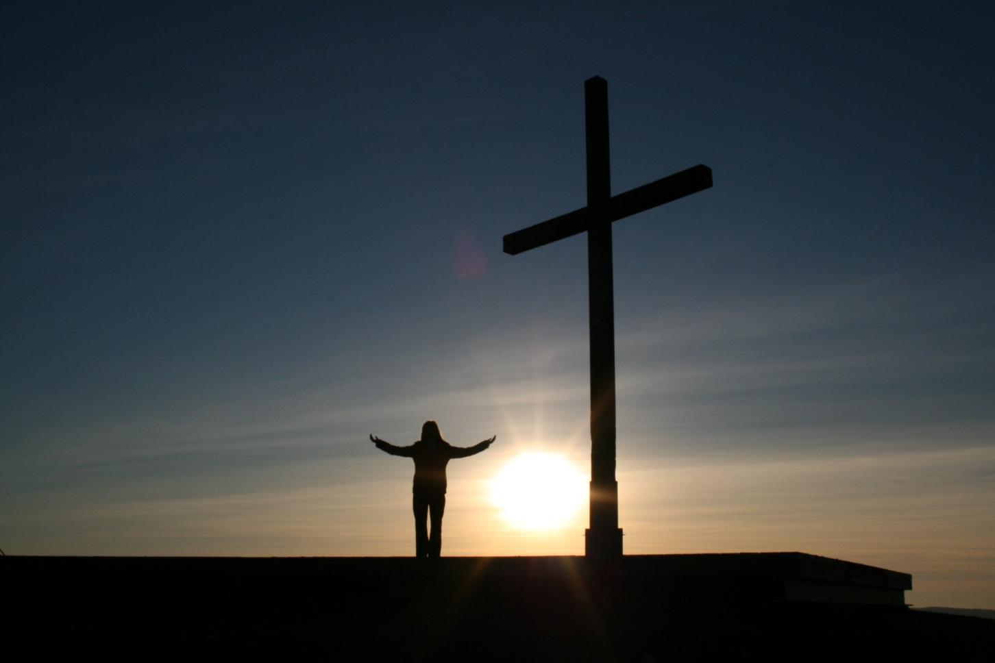 Im Vordergrund links ein Kreuz vor untergehender Abendsonne - rechts ein Mensch im Gebet mit ausgebreiteten Händen