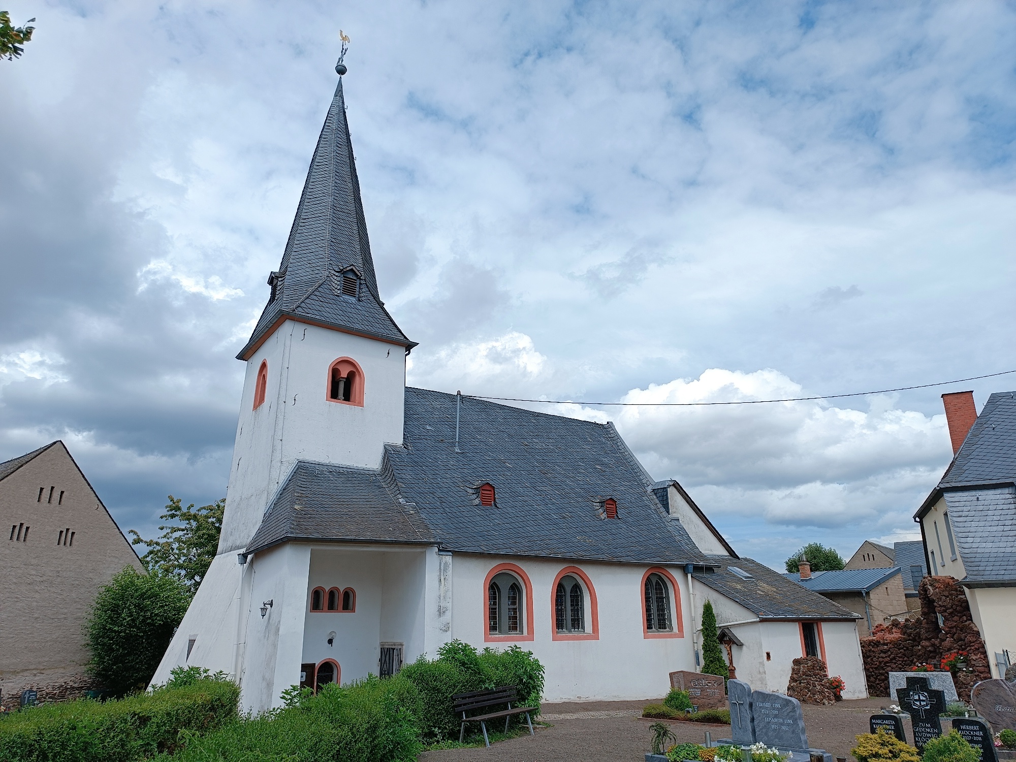 Weiße Kirchenfassade vom Friedhof aus betrachtet, links aus Sandstein gemauerte Mariengrotte