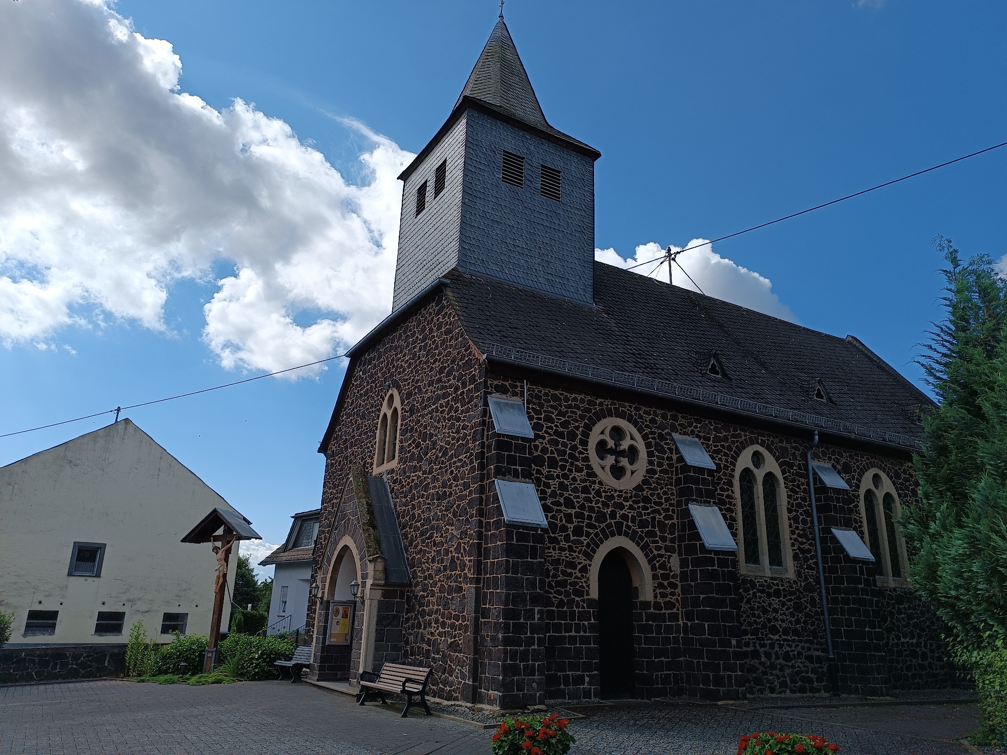 Man sieht einen dunklen Sandsteinbau mit Holzkreuz davor