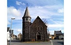 Man blickt auf das Hauptportal der Kirche, die mitten im Dorf an einer Kreuzung steht und den Kirchturm links