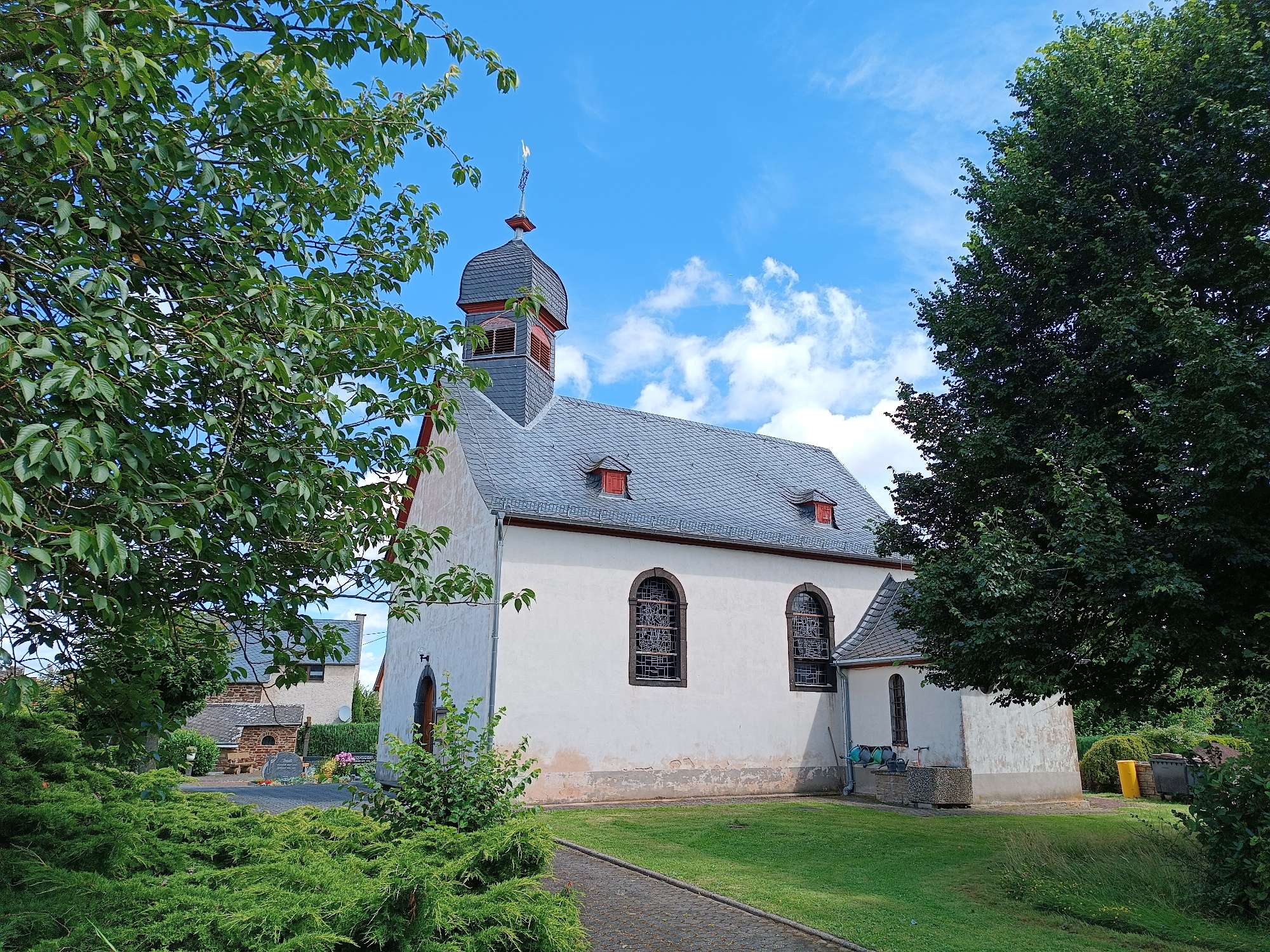 Man sieht die weiße Kirchenfassade mit Turm im Vordergrund vom Friedhof aus