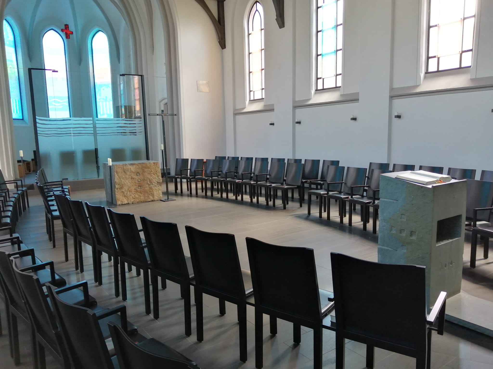 Man sieht den weiß gehaltenen Kirchenraum mit Altar links und schwarzen Stühlen rechts und links in Reihe davon