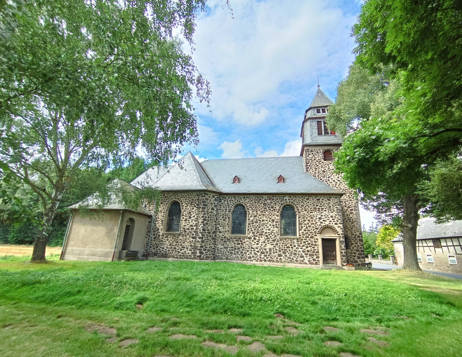 Seitenansicht der aus dunklem Sandstein gebauten Kirche bei blauem Himmel umrahmt von zwei großen Bäumen rechts und links im Vordergrund eine Wiese