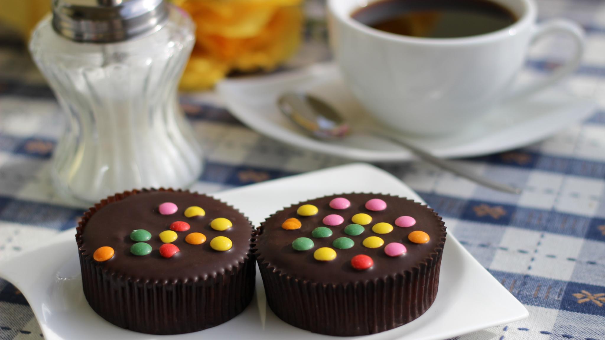 Man sieht ein weißes Kaffeegedeck mit zwei von bunten Schokolinsen bedeckten Schokoladenörtchen